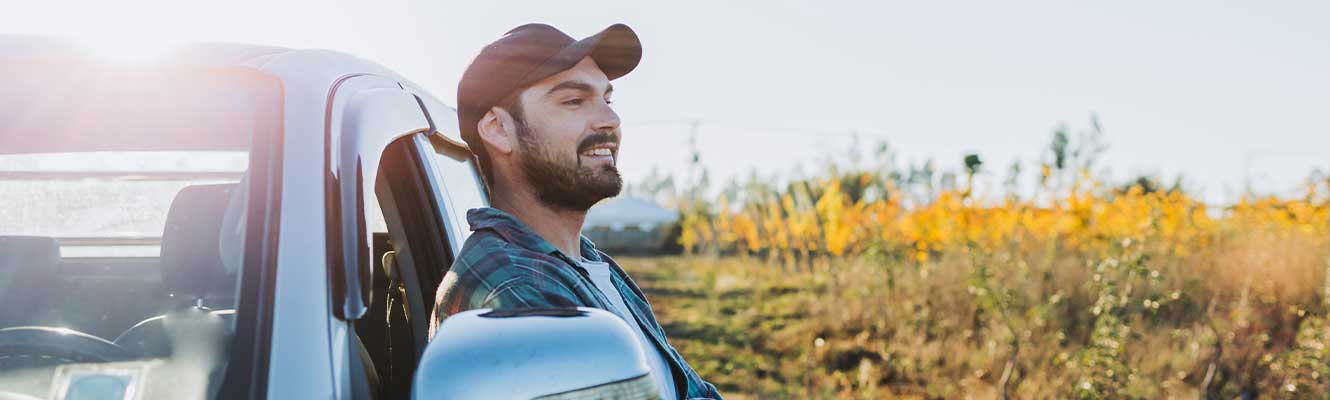Man with a ball cap leaning back on vehicle while smiling at a grassy area. 