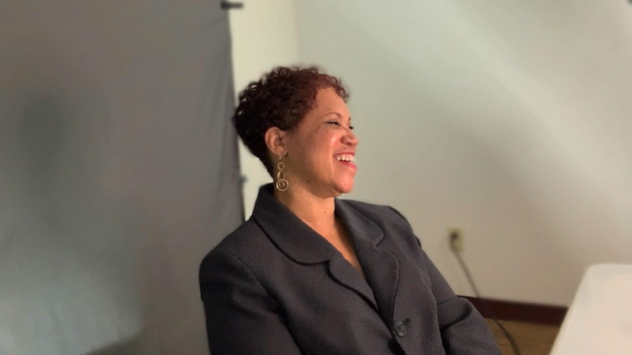 an angled shot of Carolyn Jordan looking at a different camera and laughing. She's sitting at a table with a backdrop behind her.