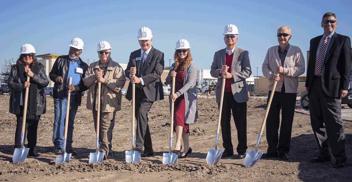 8 people lined up in a construction area with hard hats on holding shovels.