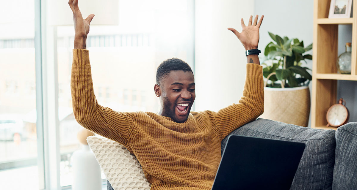 Shot of a young man cheering while using a laptop at home