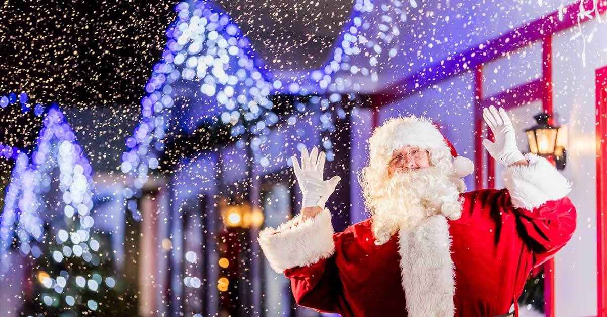 Santa Clause standing under fake snow with his hands up in the air.