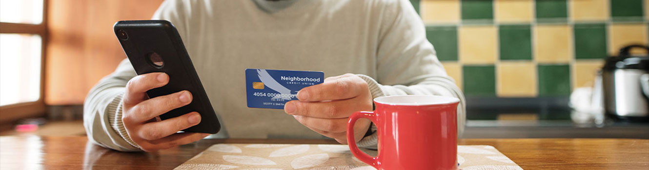 Man sitting in kitchen holding NCU debit card and looking at his phone.
