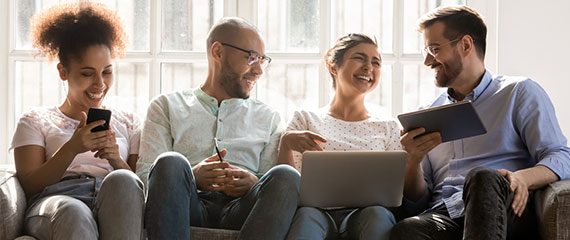 Four people sitting on a couch with books, phone, and laptop. Interacting and laughing with one another.