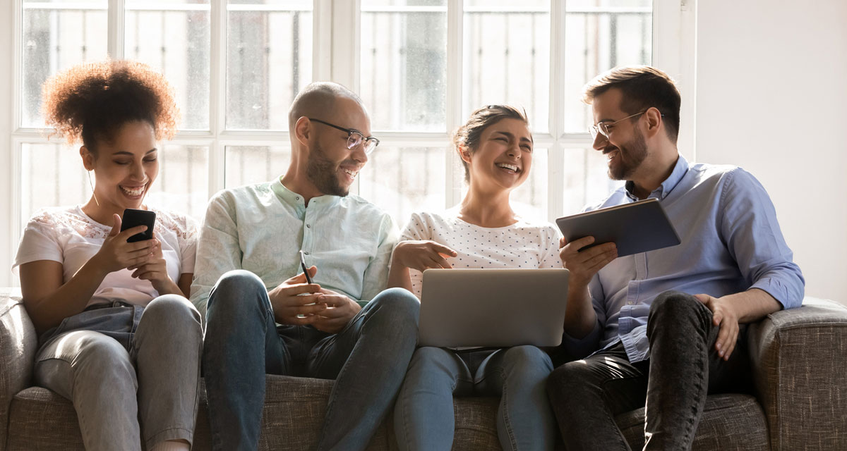 Four people sitting on a couch with books, phone, and laptop. Interacting and laughing with one another.