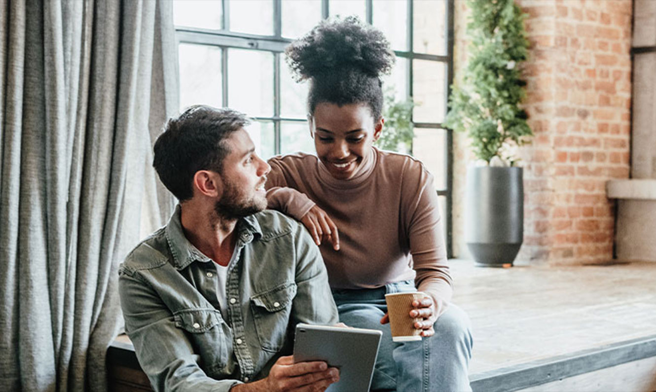 Man and woman in a loft setting are smiling and looking at an Ipad together.