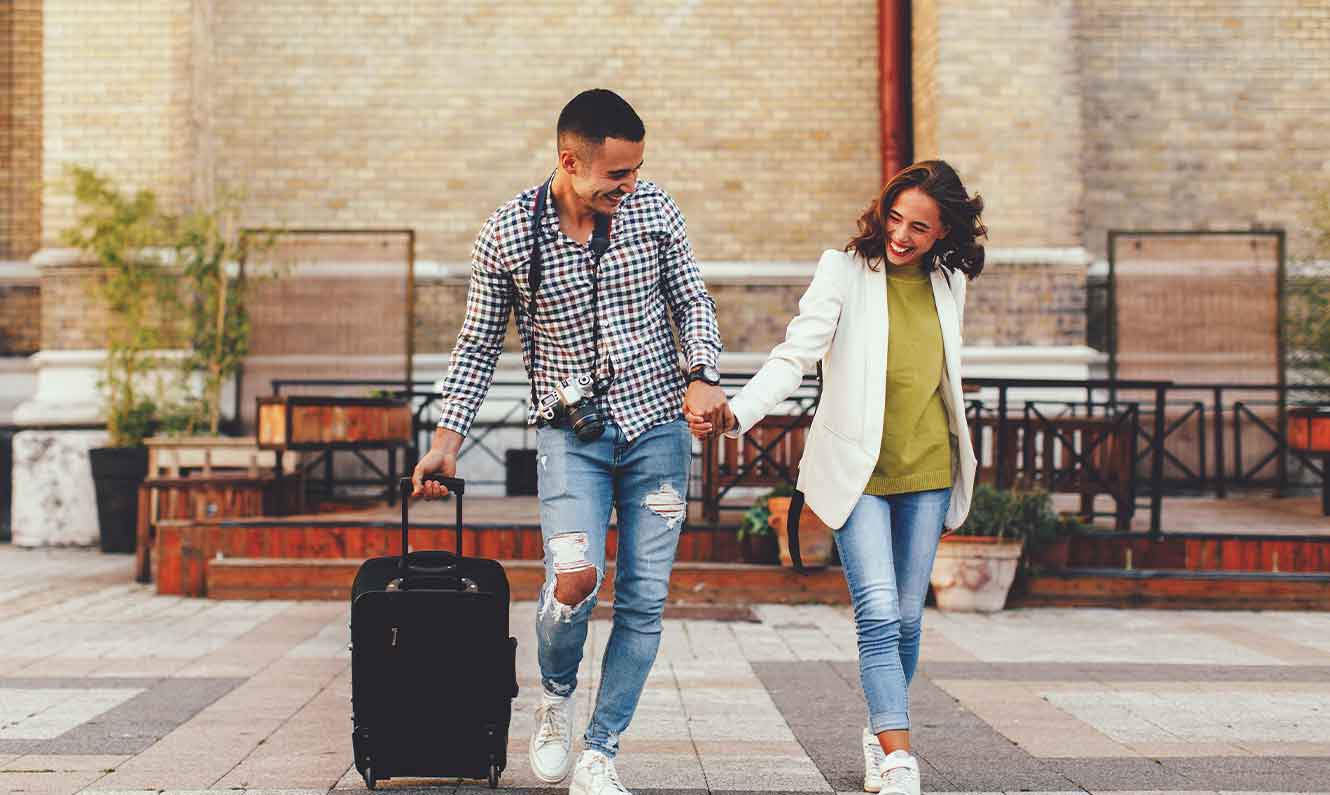 Ethnically ambiguous couple smiling holding hands and walking down a cobbled street with a suitcase.  