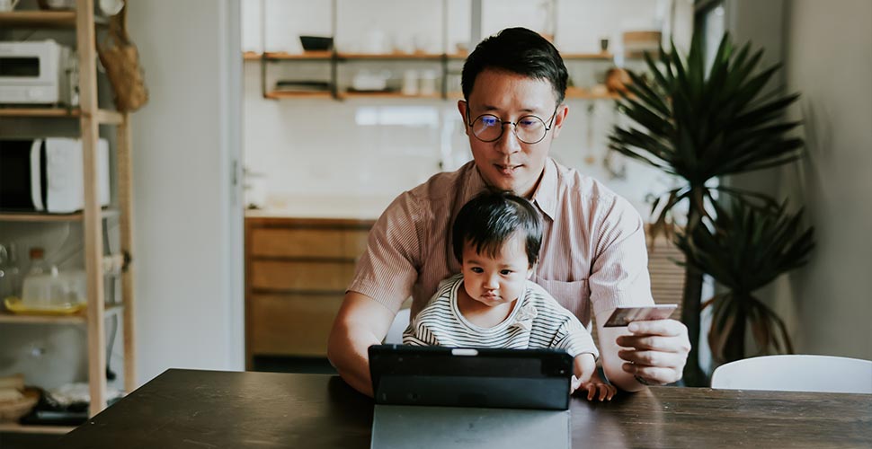 Man holding his debit card while looking at his ipad with his young child