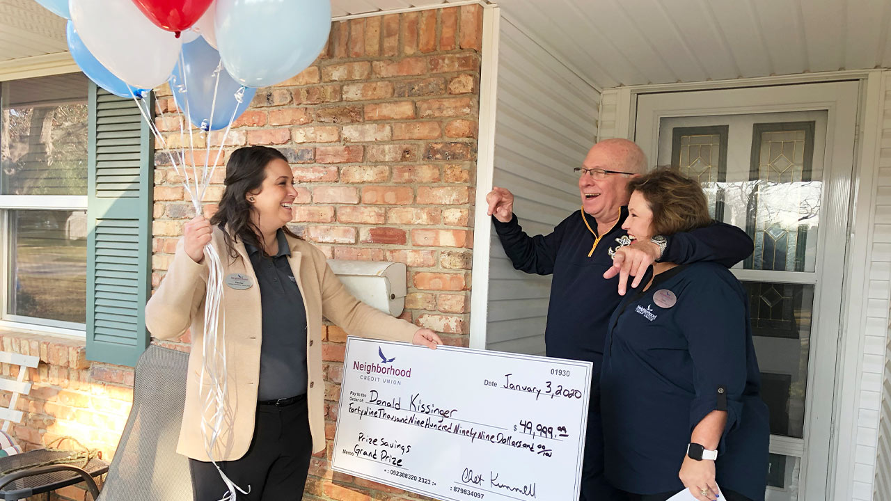 A man standing between two women. One woman is holding oversized check and balloons, other is standing by man laughing.