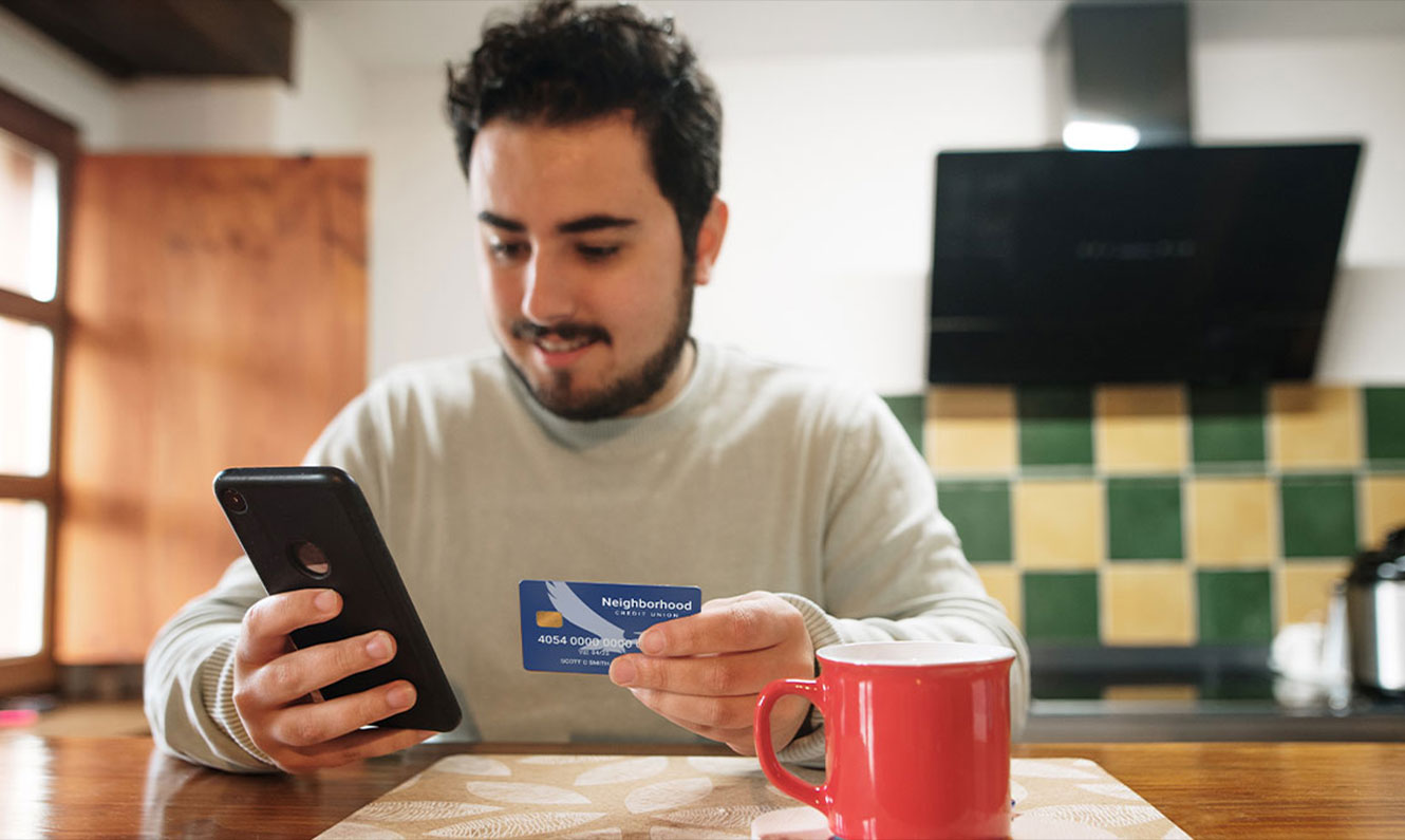 Man sitting in kitchen holding NCU debit card and looking at his phone.