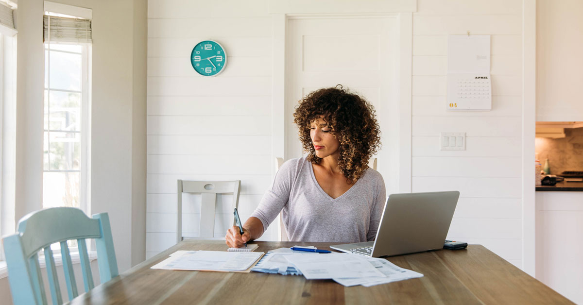 A woman sits at her dining room table with laptop and financial reports doing her taxes.