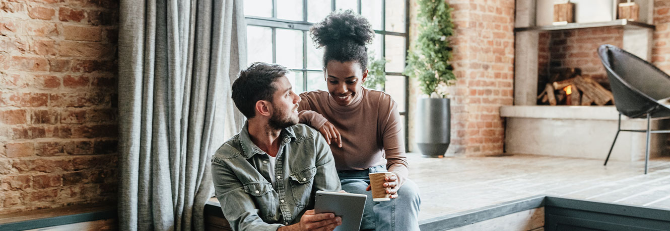 Man and woman in a loft setting are smiling and looking at an Ipad together.