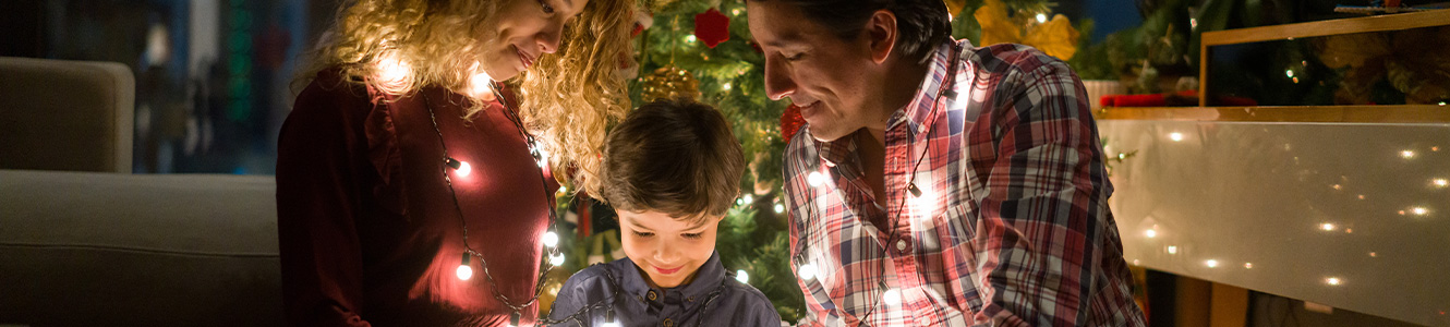 Still image of little boy with mother and father wrapped in Christmas lights sitting on the floor smiling at an open gift box.
