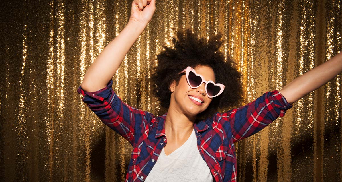 Woman dancing with heart-shaped sunglasses on in front of a gold sparkle background