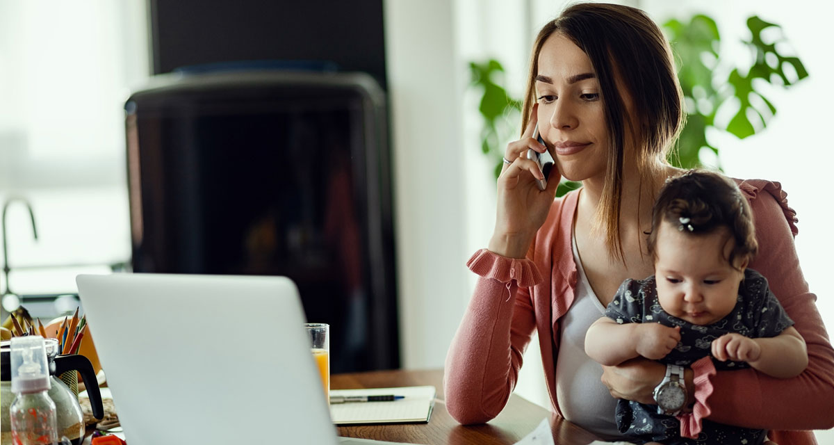 woman holding her baby while talking on the phone and looking at a laptop that is open in front of her