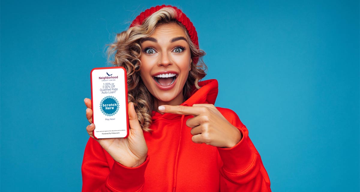Image of a woman in a bright colored hoodie looking pleasantly surprised while pointing at her phone that has the Neighborhood CU scratcher game up